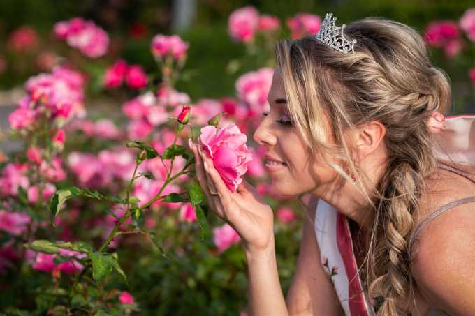 Rosenkönigin im Europa-Rosarium Sangerhausen – junge Frau mit Krone riecht an einer blühenden rosa Rose im Garten