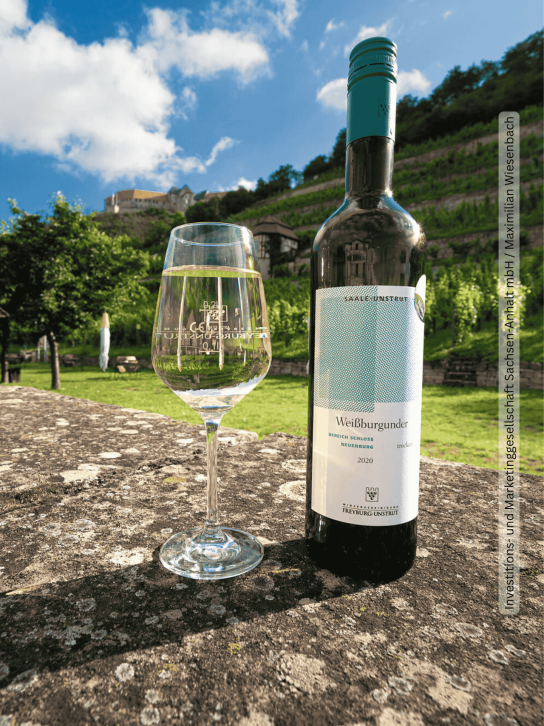 Weinglas und Flasche Weißburgunder beim Winzerfest in Freyburg, umgeben von Weinbergen mit Schloss Neuenburg im Hintergrund.